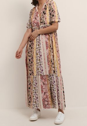 Femme portant une longue robe patchwork avec des motifs floraux et cachemire mélangés en rose, jaune et noir, assortie à des baskets blanches.