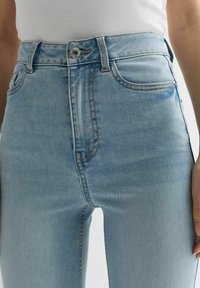 Detailní záběr na světle modré džíny s vysokým pasem, se zapínáním na knoflík a předními kapsami, nošené s bílým přízkovým topem.