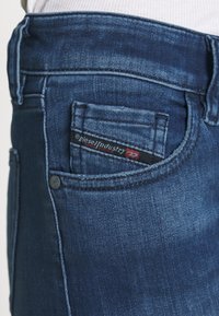 Pantalones vaqueros de mezclilla azul con un bolsillo trasero que tiene una etiqueta negra y roja, textura suave y diseño ajustado.