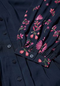 Blusa navy con stampa floreale in rosa e verde, caratterizzata da chiusure a bottone e polsini arricciati. Tessuto morbido con dettagli di design a contrasto.