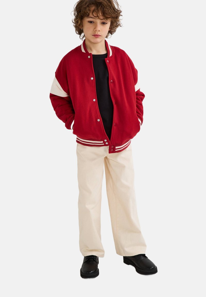 Veste bomber rouge avec des manches blanches accentuées, boutons-pression argentés, associée à une chemise noire, un pantalon crème et des chaussures noires.