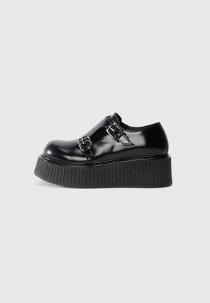 Zapato de plataforma negro brillante con suela gruesa texturizada y dos correas con hebillas plateadas a los lados, mostrado en perfil sobre un fondo blanco.
