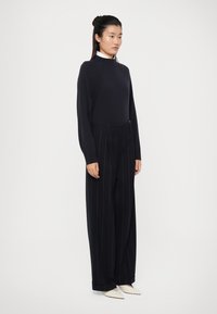Maglione a maglia navy con maniche lunghe, abbinato a pantaloni a gamba larga neri con righe sottili. Scarpe bianche a punta completano l'outfit.