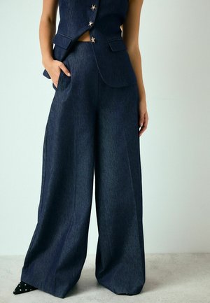 Frau trägt dunkle Jeans-Weitbein-Hose und eine passende Weste mit sternförmigen Knöpfen, Hand in der Tasche, steht auf einem hellen Boden.