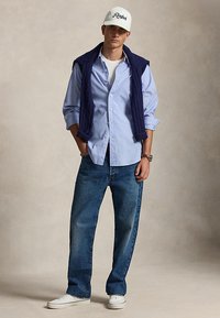 Άνδρας που φοράει λευκό καπέλο Ralph, ανοιχτό μπλε πουκάμισο με κουμπιά, σκούρο μπλε πουλόβερ ριγμένο στους ώμους, μπλε τζιν και λευκά αθλητικά παπούτσια.
