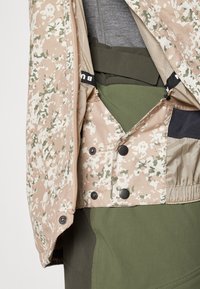 Veste camouflage avec un motif beige et vert, dotée d'une fermeture éclair et de boutons-pression noirs ; superposée à un haut gris en dessous.