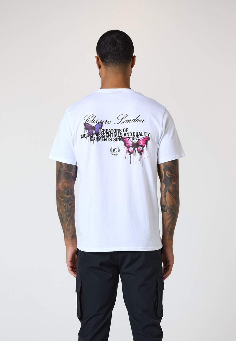 Weißes Baumwoll-T-Shirt mit buntem Schmetterlingsdruck und Text auf der Rückseite, kombiniert mit schwarzen Cargo-Hosen. Schlichtes und lässiges Design.