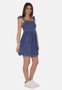 Abito estivo in denim con spalline arricciate, corpetto aderente e gonna svasata. Tessuto blu scuro con cuciture a contrasto, indossato con sneaker bianche.