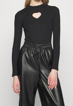 Vrouw draagt een zwarte geribde top met hartvormige uitsparing op de borst en zwarte nepleren broek met hoge taille en trekkoord in de taille.