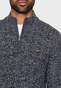 Gestrickter Reißverschluss-Pullover in einem schwarz-weißen Strukturmuster, mit einem kleinen gelben Logo auf der Brust und einem hohen Kragen.