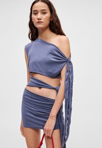 Σετ που αποτελείται από τοπ ενός ώμου, κοντό, και μίνι φούστα σε απλό ανοιχτό μπλε ύφασμα. Διαθέτει στριφογυρισμένους ιμάντες και κόμπο στη μέση.