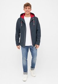 Navy jacket with red hood and accents, featuring a zip front, two side pockets, and drawstrings. Paired with light blue jeans and white sneakers.