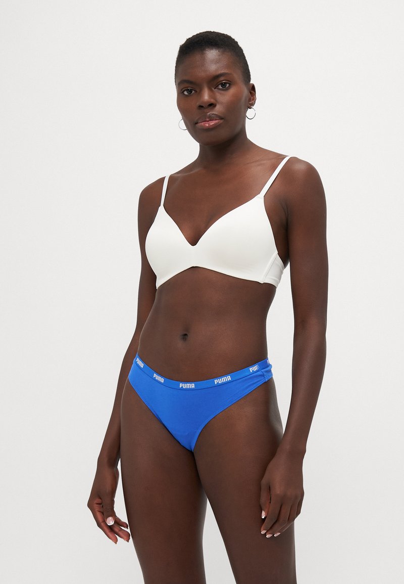Vit, sömlös bralette med tunna remmar i kombination med blå underkläder som har en Puma-logotyp på midjebandet. Slät textur, samtida design.