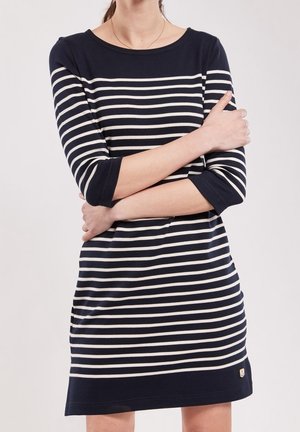 Woman wearing a navy blue and white horizontal striped dress with three-quarter sleeves, arms crossed over chest.
