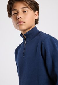 Laivastonsininen vetoketjullinen neulehuppari, jossa on korkea kaulukko, teksturoidusta neuleesta valmistettu kangas ja hopeinen vetoketju, soveltuu rentoon käyttöön.