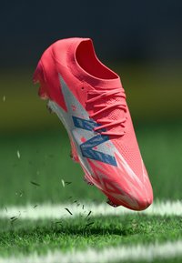 Svetlo roza nogometna kopačka z teksturirano zgornjo površino, belimi in modrimi detajli ter aerodinamičnim dizajnom, viseča nad zeleno travo.