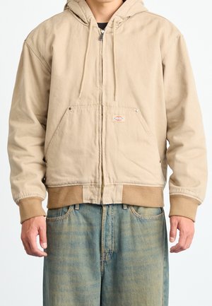 Mand iført beige hættejakke med lynlås, forlommer og manchetter, kombineret med lyseblå jeans.
