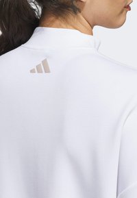 White athletic shirt featuring a high collar, smooth fabric texture, and a subtle beige three-stripe logo on the back.
