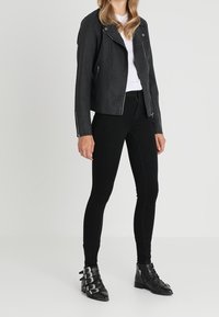 Chaqueta de motociclista de cuero sintético negro con cremallera asimétrica y herrajes plateados, combinada con jeans ajustados negros y botines al tobillo.