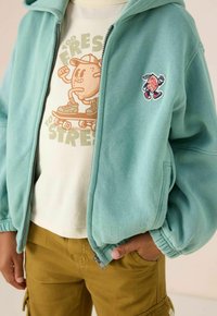 Hoodie zippé vert menthe avec des poignets élastiques, comportant une patch multicolore. Associé à un t-shirt graphique beige et un pantalon cargo moutarde.