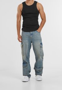 Černé tílko spojené s lehce modrými volnými cargo džínami s mnoha kapsami a vybledlým designem, nošené s bílými teniskami.