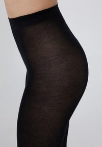 Collants opaques noirs avec une texture lisse, présentant une taille haute et une coupe ajustée, vus de côté.