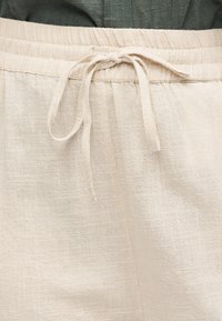 Μπεζ παντελόνι λινό με ελαστική μέση και δεμένο κορδόνι, φορεμένο με σκούρο πράσινο τοπ.