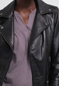 Giacca da motociclista in pelle nera con colletto a revers, chiusura frontale con zip e dettagli in argento, indossata sopra una maglia viola chiaro con scollo a V.