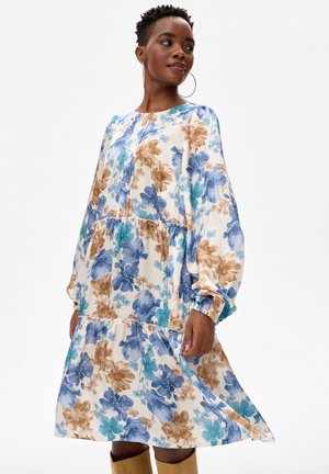 Kobieta w niebiesko-turkusowo-brązowej, kwiecistej sukience o długości do kolan z długim rękawem, w parze z beżowymi butami, na jednolitym białym tle.