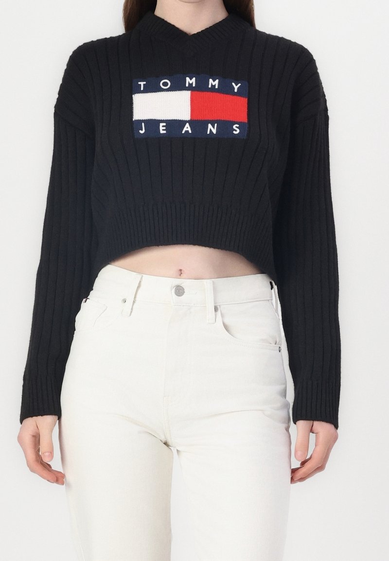 Czarny, prążkowany sweter krótki z dekoltem w kształcie litery V i naszywką z logo z przodu w kolorach czerwonym, białym i niebieskim. Zestawiony z białymi, wysokim stanem dżinsami.