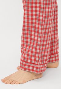 Rode geruite pyjamabroek van zacht materiaal, met een klassiek ruitpatroon in wit en zwart en opgestikte manchetten. Blote voet zichtbaar.