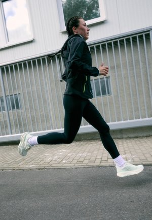 Running shoes in light green with textured sole, paired with black leggings and a black jacket. The runner is in mid-stride on a paved surface.