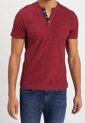 Rode korte mouw Henley shirt van een gestructureerde stof, met een knoopsluiting en contrasterende halslijn. Draagt met blauwe jeans.