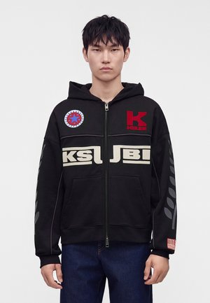 Jeune homme portant un sweat à capuche noir zippé avec des patchs rouges et blancs et des logos, debout devant un fond blanc uni.