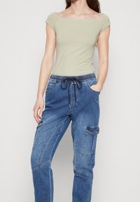Kvinna som bär en ljusgrön off-shoulder kortärmad topp och blå denim cargo-byxor med elastisk midja och dragsko, stående med händerna i fickorna.