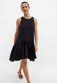 Mustat olkaimettomat mekko, jossa on teksturoitu pinta ja röyhelöhelma. Suunnittelussa on epäsymmetrinen leikkaus ja rento istuvuus.