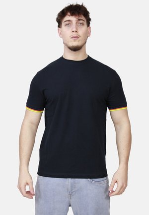 Giovane uomo con t-shirt nera con rifiniture rosse e gialle sulle maniche e jeans azzurri chiari, in piedi davanti a uno sfondo bianco.