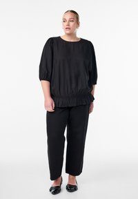 Melnā blūze ar pūkainām piedurknēm, savilktu jostasvietu un frēzētu apakšmalu, apvienota ar melnām taisnām biksēm un spīdīgām melnām kurpēm.