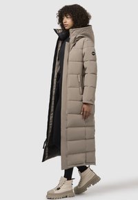Abrigo largo de plumas beige con un collar alto, costuras horizontales y bolsillos con solapa. Llevado sobre un conjunto negro, combinado con botas gruesas beige.