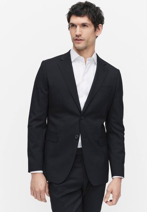 Man draagt een zwarte tweeknops colbert en bijpassende broek met een wit overhemd, staand tegen een effen witte achtergrond.