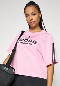 Kvinne med flettet hår iført en rosa Adidas treningsskjorte og svart bærestropp over skulderen, som står foran en ensfarget bakgrunn.