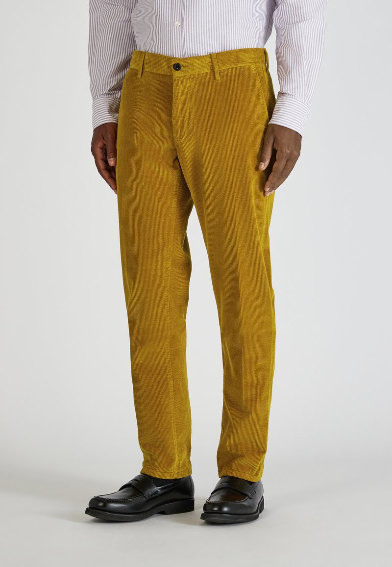 Geel corduroy broek met verticale ribbels, voorzien van een rits en knoopsluiting aan de voorkant, gecombineerd met zwarte schoenen.