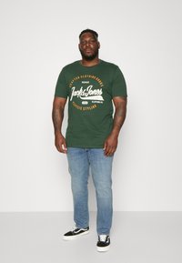 Mørkegrønn bomulls t-skjorte med hvit og oransje grafisk tekst, kombinert med lyseblå denimjeans og svart-hvite sneakers.