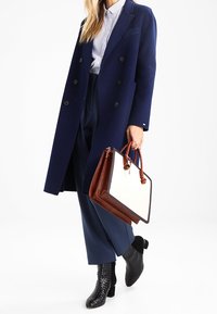 Granatowy wełniany płaszcz dwurzędowy nałożony na jasną koszulę w paski, połączony z granatowymi spodniami i czarnymi teksturowanymi botkami za kostkę, trzymający torebkę w dwóch kolorach.