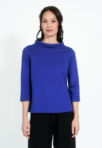 Kobalt kék cowl-nyakú felső, puha anyagból készült. Háromnegyedes ujjakkal és laza szabással rendelkezik. Fekete nadrággal viselve.