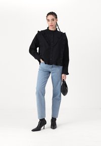 Svart rufset knappejakke med høy hals, parret med lyseblå rett-bein jeans og svarte spisse ankelstøvler, holder en svart veske.