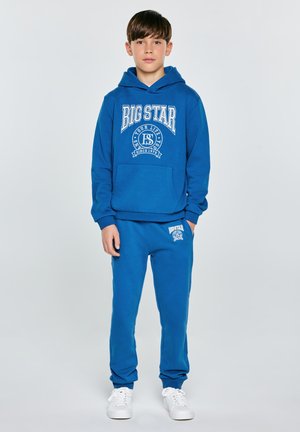 Chlapec stojí, oblečený v modré mikině Big Star s kapucí a odpovídajících teplácích, s bílými teniskami, na jednoduchém světle šedém pozadí.
