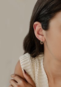 Pendientes de aro de oro con un diseño retorcido, que presentan un acabado brillante. Llevados por un modelo con cabello largo y oscuro y una blusa blanca texturizada.