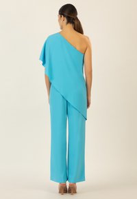 Jumpsuit turchese a spalla unica con drappeggio fluido, pantaloni a gamba larga e superficie liscia. La vista posteriore mette in mostra un design pulito e hardware minimale.