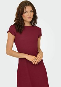Robe ajustée bordeaux avec des manches courtes et retroussées. Tissu texturé et encolure ronde. Longueur sous le genou avec une silhouette lisse et épurée.